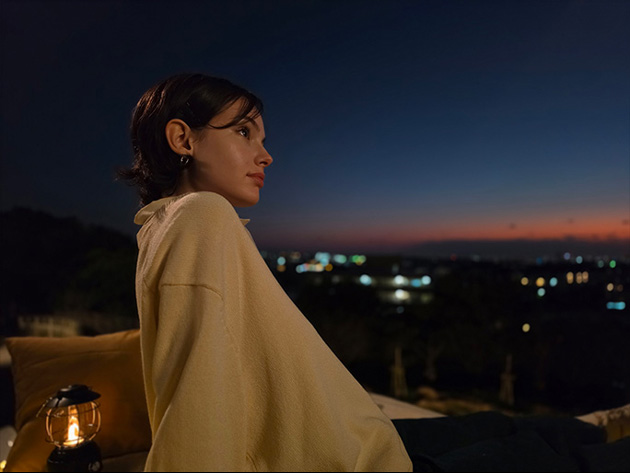 Low-light portrait of a person looking at a city skyline at dusk. The image highlights the S26 Ultra’s ability to capture sharp skin textures and fabric details under dim conditions, with a high-contrast background of city lights and a deep blue twilight sky.