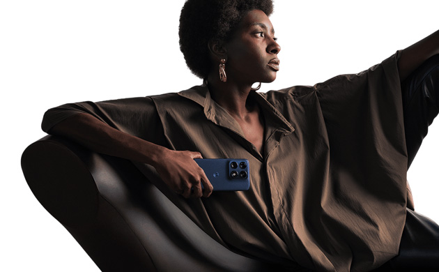 A Black woman with natural hair, wearing a loose-fitting brown top and large gold earrings, is seated and holding a Titan color of Motorola Edge 70 Pro.
