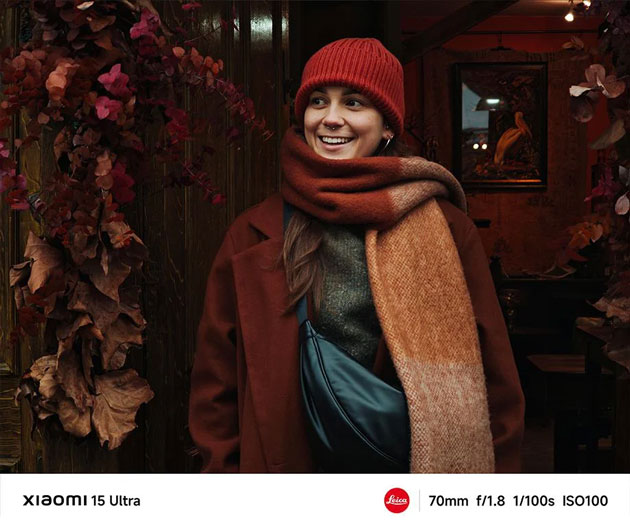 A cozy outdoor portrait captured with the Xiaomi 15 Ultras 70mm telephoto lens at f1.8 A cozy outdoor portrait captured with the Xiaomi 15 Ultra's 70mm telephoto lens at f/1.8. A smiling woman wearing a red beanie, a thick patterned scarf, and a burgundy coat stands next to a cluster of dried autumn leaves.