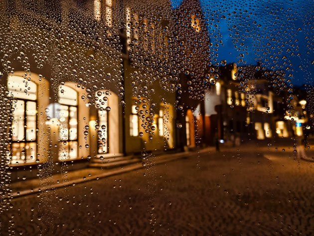A low-light camera sample from the Realme C71 showing water droplets on a glass surface with a blurred European-style city street and warm building lights in the background at night.