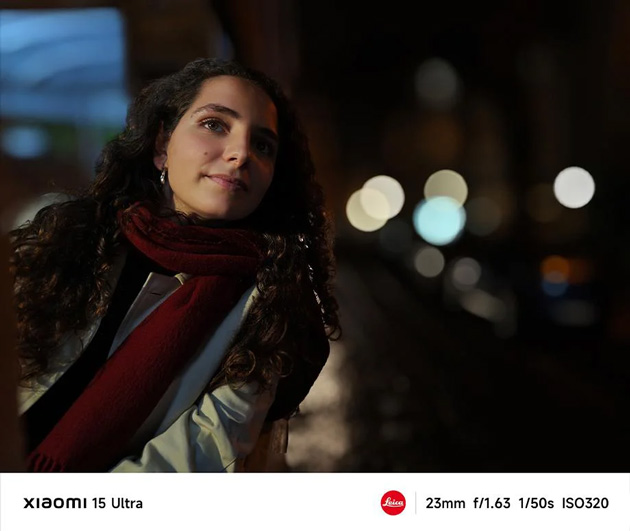 A low light portrait captured with the Xiaomi 15 Ultras 23mm main lens at f1.63 A low-light portrait captured with the Xiaomi 15 Ultra's 23mm main lens at f/1.63. The photo features a woman with long curly hair and a red scarf, with her face softly illuminated against a dark background.