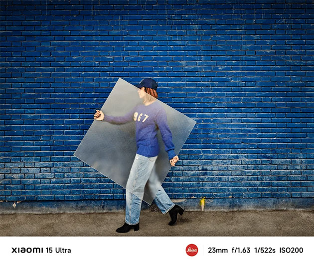 A vibrant street photograph captured with the Xiaomi 15 Ultras 23mm main lens A vibrant street photograph captured with the Xiaomi 15 Ultra's 23mm main lens. A person in a blue sweater and jeans walks past a bright blue brick wall while carrying a large, semi-transparent frosted square.