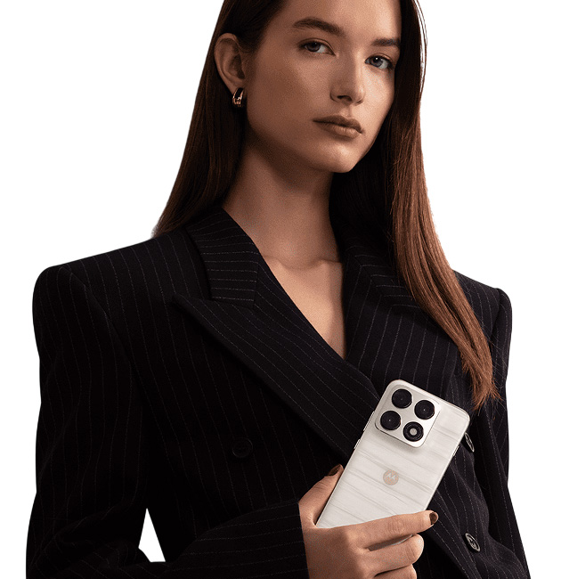 A woman with long, dark brown hair, wearing a black pinstripe blazer, holds a Lily White Motorola Edge 70 Pro.