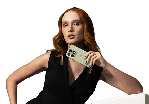 A woman with long, wavy auburn hair, wearing a black sleeveless dress, holds a Tea color of the Motorola Edge 70 Pro.