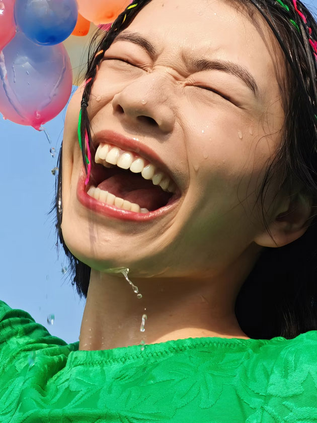 Portrait shot taken on the Honor 400 Pro showing a young woman laughing joyfully with her eyes closed while being splashed by colorful water balloons against a clear blue sky Portrait shot taken on the Honor 400 Pro, showing a young woman laughing joyfully with her eyes closed while being splashed by colorful water balloons against a clear blue sky.