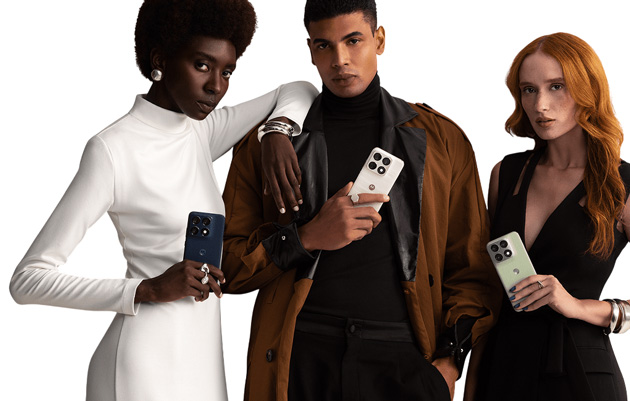 Three models, two women and one man, pose with Motorola Edge 70 Pro phones. A Black woman in a white dress holds a dark blue (Titan) phone, a man in a brown jacket holds a Lily White phone, and a woman with red hair in a black dress holds a light green (Tea) phone. All have plain white backgrounds.