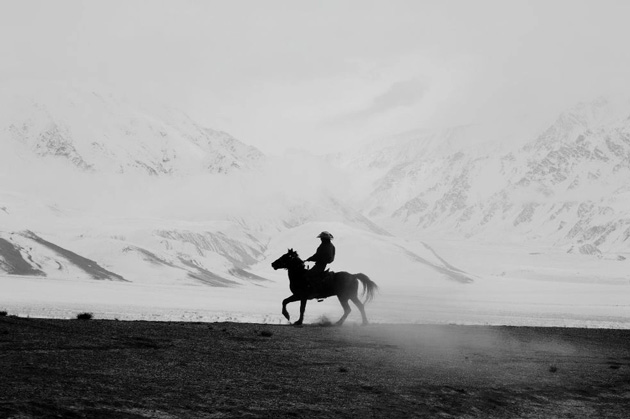 Black-and-white photo of a horseback rider in nature, with snowy mountains and scattered fog in the background, wide frame with shallow detail; photo taken with OPPO Find X9 Ultra camera