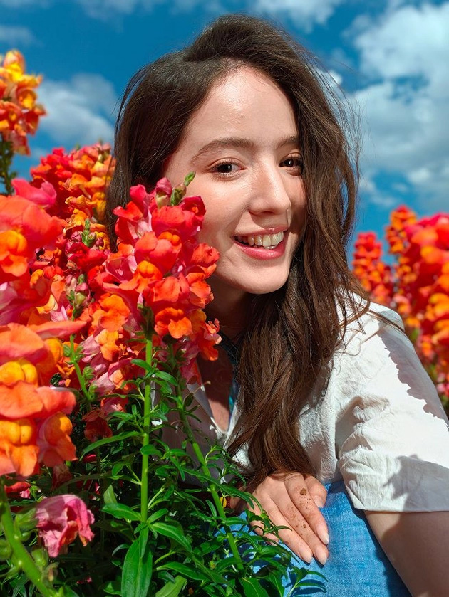 Close-up portrait outdoors next to colorful bouquets, blue sky with scattered clouds and a blurred background; photo taken with OPPO Find X9 Ultra camera