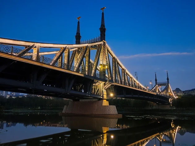 Nighttime photo sample from the Realme C75 showing a grand illuminated bridge with European style spires reflecting in the calm water below against a deep blue twilight sky Nighttime photo sample from the Realme C75 showing a grand illuminated bridge with European-style spires, reflecting in the calm water below against a deep blue twilight sky.