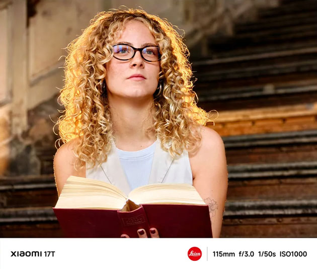 A portrait photo taken with the Xiaomi 17T's camera, featuring a woman with curly hair and glasses who's reading a book on the stairs.