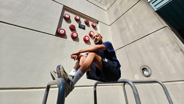 Photo taken with the Nothing Phone 3 ultrawide camera of a person sitting on metal bars in front of a wall with red bells Photo taken with the Nothing Phone (3) ultrawide camera of a person sitting on metal bars in front of a wall with red bells.